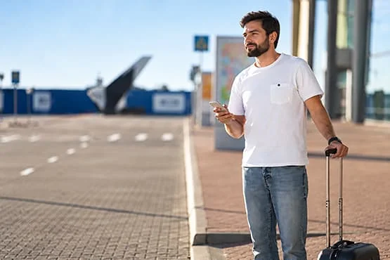 Un voyageur tient son téléphone et la poignée de sa valise à l’extérieur d’un terminal.
